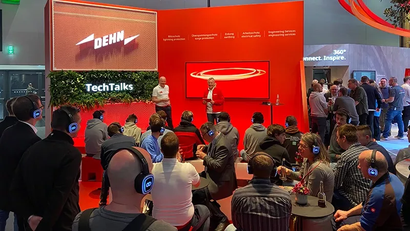 A group of people attend a TechTalks presentation at a DEHN booth; speakers stand near a screen while the audience listens with wireless headphones.