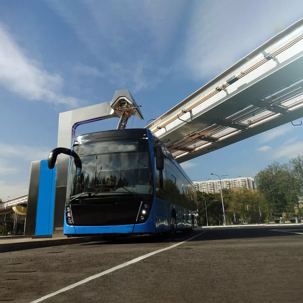 Blue electric bus at a charging station beneath an elevated pedestrian bridge.