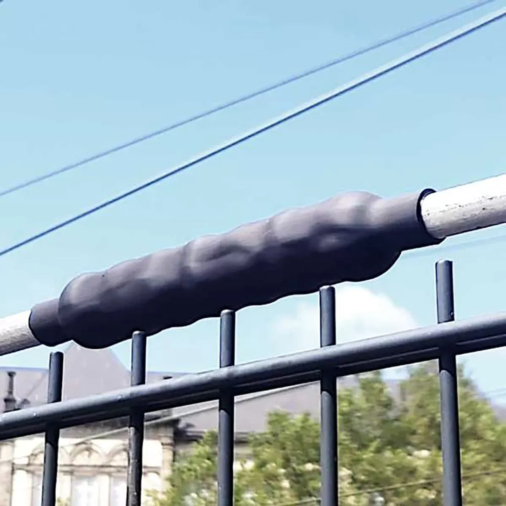 Close-up of a black protective sleeve on a metal fence bar with wires and a building in the background.