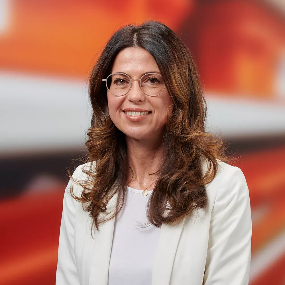 Professional portrait of a woman with glasses and shoulder-length brown hair, wearing a white blazer and smiling against a warm, abstract background.