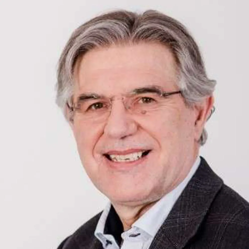 Portrait of a gray-haired man wearing glasses and a dark jacket over a light shirt, smiling against a white background.