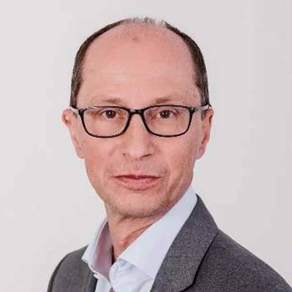 Portrait of a man wearing glasses, a gray jacket, and a light shirt against a white background.